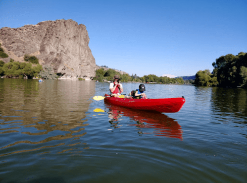A person and a child paddle a red kayak on a calm lake, surrounded by trees and rocky cliffs under a clear blue sky.