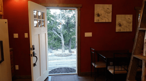 A cozy kitchen with a door open to a deck, featuring red walls, a small dining table, and nature views outside.