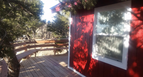 A red cabin with a wooden deck surrounded by trees, casting shadows on the ground in bright sunlight.