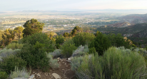 A scenic view from a mountain overlooking a valley filled with greenery and distant mountains under a clear sky.