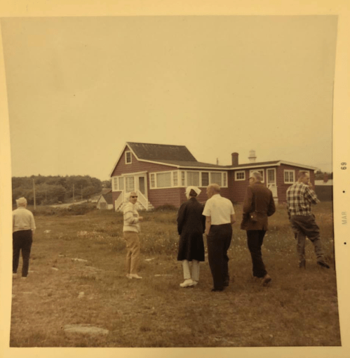 A group of people walking towards a red house in a rural setting, with grassy fields and overcast skies.