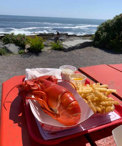 A red lobster with fries and dipping sauce on a red table, overlooking a scenic ocean view.