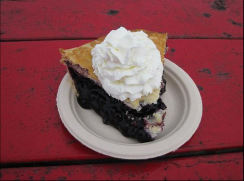 A slice of blueberry pie topped with whipped cream on a paper plate, resting on a red wooden table.
