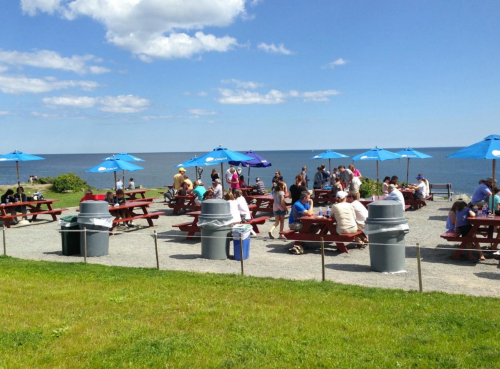A lively outdoor dining area with picnic tables and umbrellas by the ocean, filled with people enjoying the sunny day.