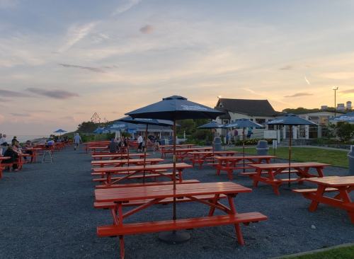 Outdoor dining area with red picnic tables and umbrellas at sunset, near a beachside restaurant.