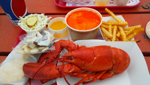 A plate featuring a whole red lobster, fries, coleslaw, a cup of soup, and condiments on a red tray.