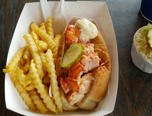 A lobster roll with mayonnaise and pickles, served with crinkle-cut fries in a takeout container.
