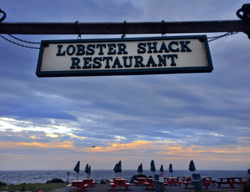 Sign for "Lobster Shack Restaurant" with picnic tables and ocean view under a cloudy sky at sunset.