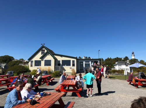 A busy outdoor dining area with red picnic tables, people enjoying food, and a lighthouse in the background under a clear blue sky.