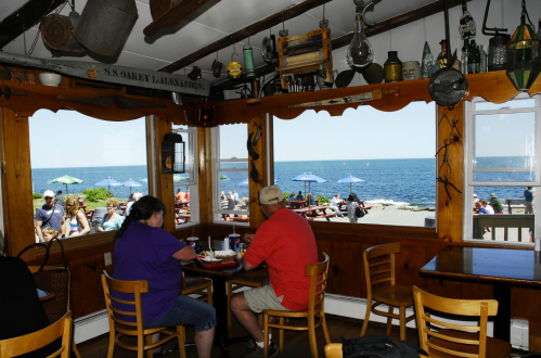 Two people sit at a table inside a seaside restaurant, with a view of the ocean and outdoor seating visible.