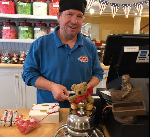 A smiling man in a blue shirt and black hat stands at a candy shop counter with a teddy bear and treats.