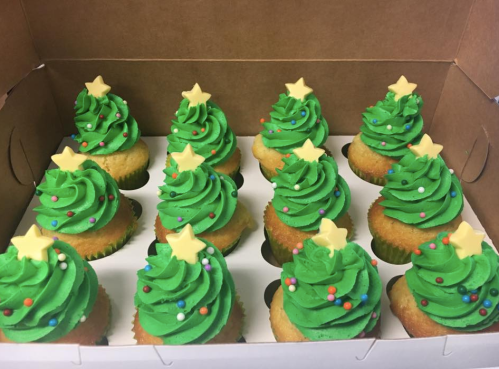 A box of festive cupcakes decorated as Christmas trees, topped with green frosting, colorful sprinkles, and yellow stars.