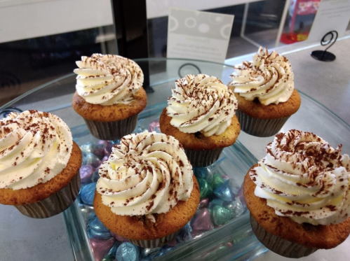 A display of six cupcakes with swirled frosting, topped with cocoa powder, on a glass platter with colorful candy.