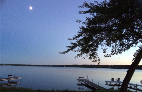A serene lake at dusk, with a crescent moon and silhouettes of trees and docks along the water's edge.