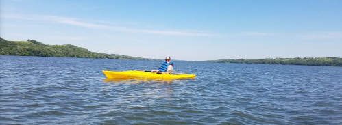 A person in a blue life jacket paddles a yellow kayak on a calm lake under a clear blue sky.