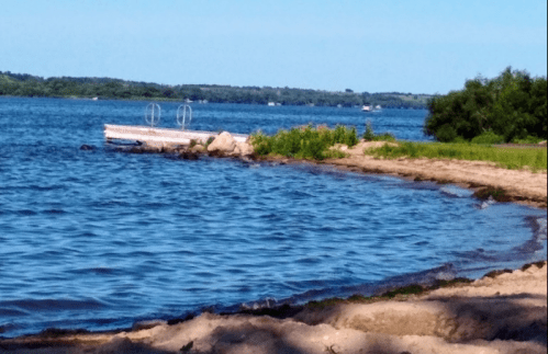 A serene lakeside view with a sandy shore, a small dock, and lush greenery under a clear blue sky.