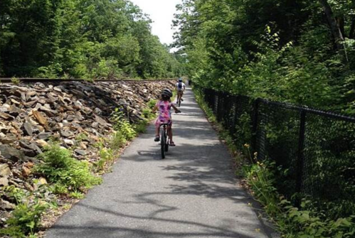 A child rides a bike on a path beside a stone wall, surrounded by lush greenery and trees.