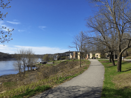 A scenic riverside path lined with trees, leading to houses under a clear blue sky.