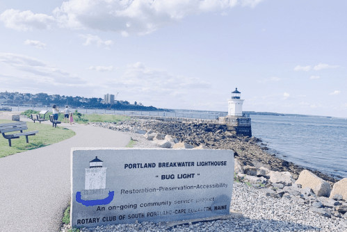 A scenic view of Portland Breakwater Lighthouse with a sign, walking path, and people enjoying the waterfront in Maine.