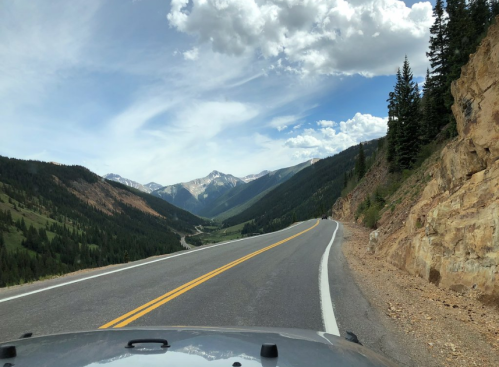 A winding mountain road with lush greenery and rocky cliffs, under a partly cloudy sky.