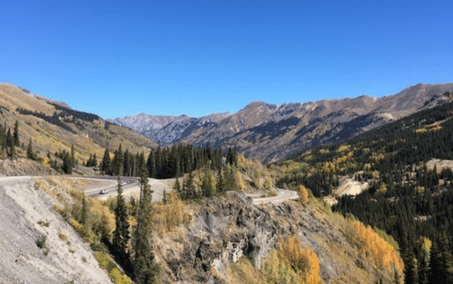Scenic mountain landscape with winding road, vibrant autumn foliage, and clear blue sky.