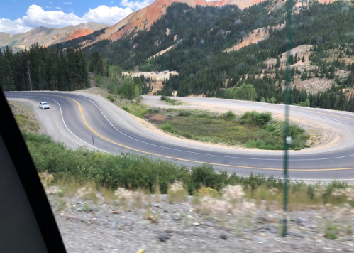 A winding road curves through a mountainous landscape with trees and colorful rock formations in the background.