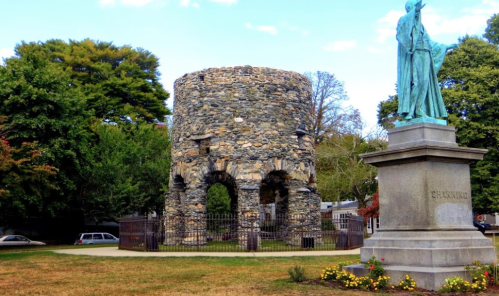 A stone tower ruins surrounded by greenery, with a statue nearby and flowers in the foreground.