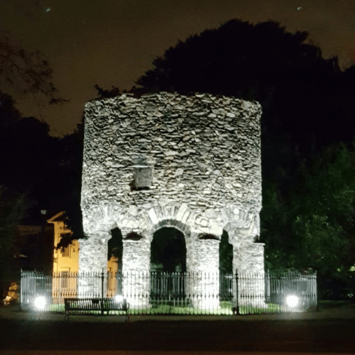 A stone structure illuminated at night, surrounded by trees and a fence, with lights highlighting its features.