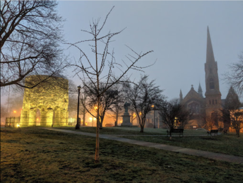 Foggy scene featuring a historic stone structure and a church steeple, illuminated by streetlights in a park.