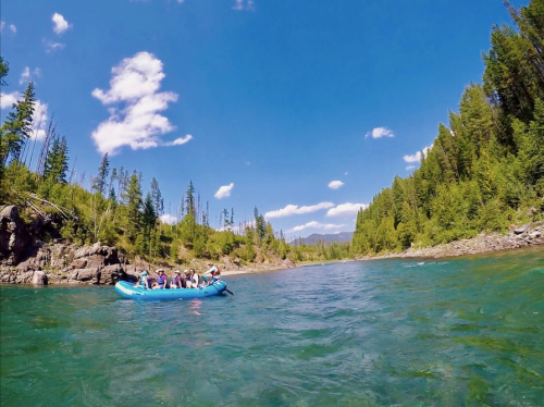 A group of people rafting on a clear river surrounded by lush green trees and a bright blue sky.