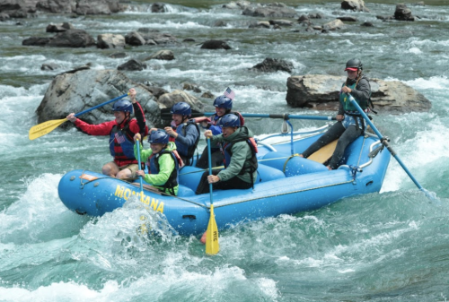 A group of six people in a blue raft navigating whitewater rapids, wearing helmets and paddling with yellow oars.