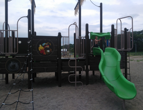 A playground with a green slide and climbing structures, set in a sandy area under a cloudy sky.