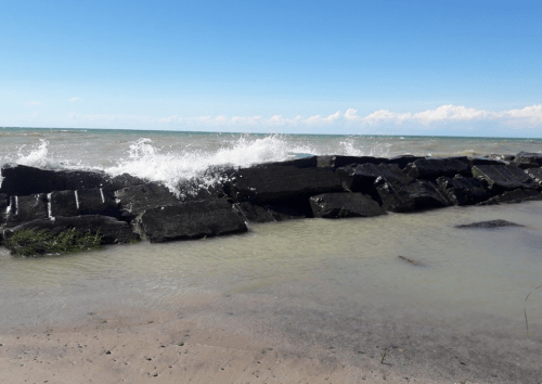 Waves crash against a rocky jetty at the beach under a clear blue sky.