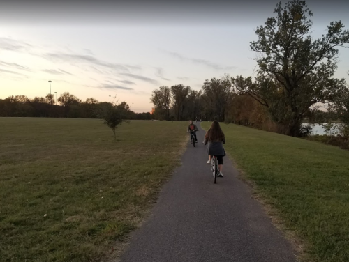 Two children ride bicycles along a path in a park during sunset, surrounded by trees and open grass.