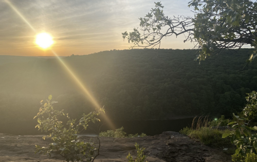 Sunset over a lush green landscape, with a river visible below and sunlight streaming through the trees.