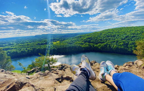 Two pairs of feet resting on a rocky ledge, overlooking a lush green landscape and a winding river under a blue sky.