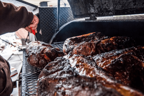 A person uses a knife to slice smoked meat on a grill, with several large cuts of meat visible.