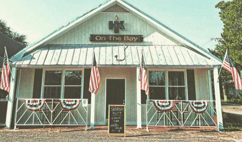A white building with a sign reading "HK On The Bay," decorated with American flags and bunting for a July 4th event.