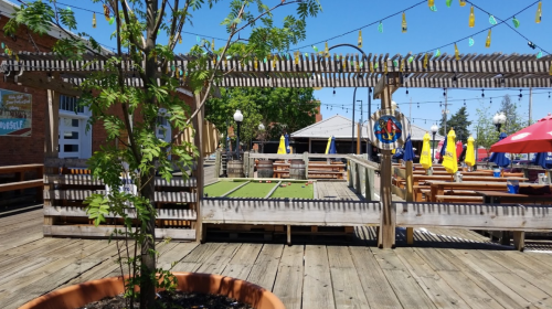Outdoor dining area with wooden decking, string lights, and colorful umbrellas, featuring a bocce ball court.
