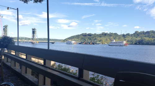 A scenic view of a river with boats, greenery on the shore, and a clear blue sky with scattered clouds.