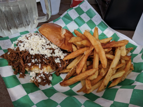 A plate with a pulled pork sandwich topped with cheese and a side of golden fries, served on a green checkered paper.