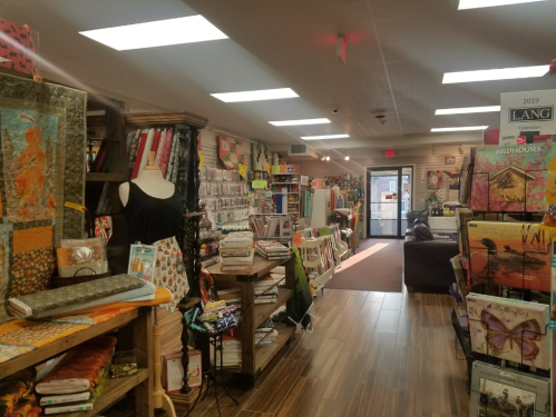 A cozy fabric store aisle filled with colorful fabrics, quilting supplies, and craft items, leading to the entrance.