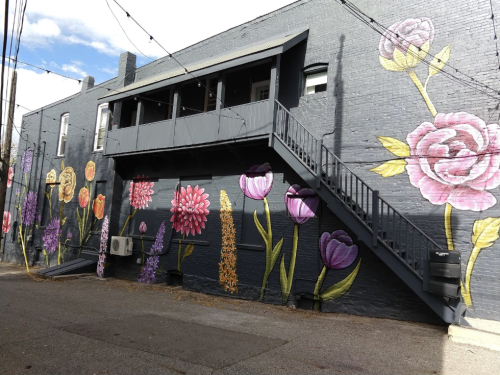 Colorful flower murals adorn a black brick wall, featuring various blooms and a staircase leading to a second-floor balcony.