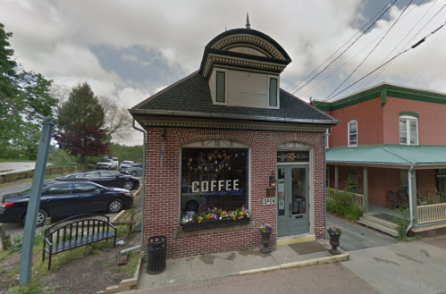 A charming brick coffee shop with a sign reading "COFFEE" and an open door, surrounded by trees and parked cars.