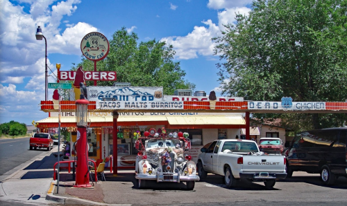 A retro diner with a colorful sign, vintage cars parked outside, and a clear blue sky with fluffy clouds.
