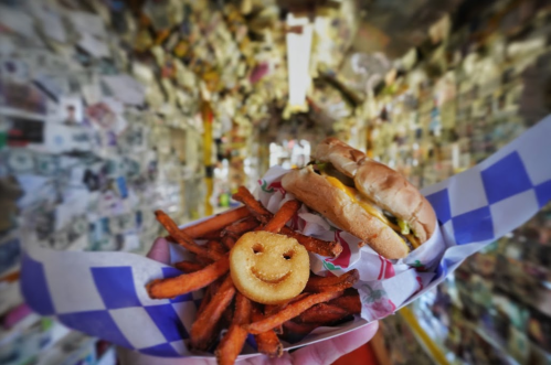 A close-up of a burger and sweet potato fries with a smiley face potato, set against a colorful, cluttered background.