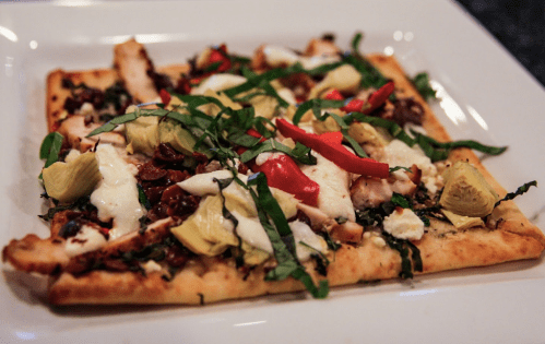 A flatbread pizza topped with chicken, artichokes, red peppers, basil, and mozzarella on a white plate.
