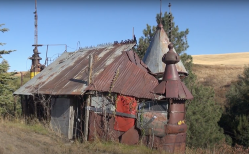 A quirky, weathered metal house with unique architectural features, surrounded by trees and rolling hills.