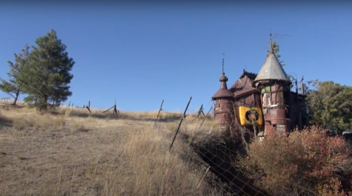 A whimsical, castle-like structure sits on a grassy hillside, surrounded by a fence and trees under a clear blue sky.