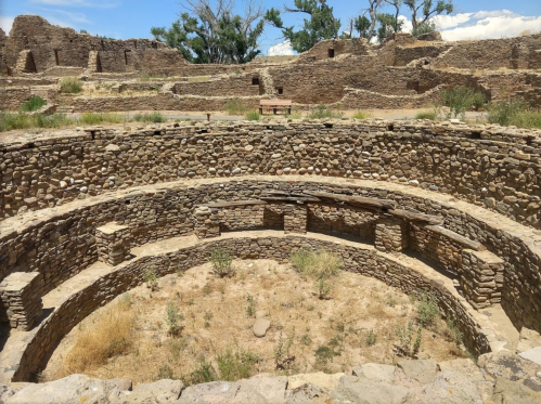 Ancient stone ruins with circular structure and grassy areas under a blue sky with scattered clouds.